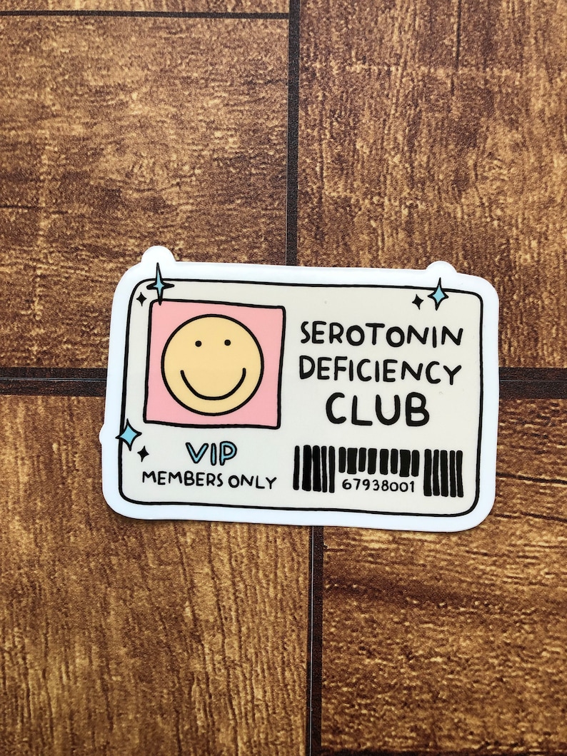 May include: A white sticker with a cartoon smiley face, the text "SEROTONIN DEFICIENCY CLUB", "VIP MEMBERS ONLY", and a barcode with the number "67938001".