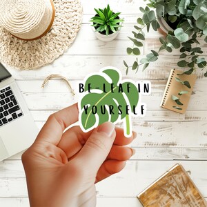 May include: A die-cut vinyl sticker shaped like a green monstera leaf with the black text BE-LEAF IN YOURSELF printed across the center. The sticker features a white border and a flat, matte finish, showcasing a stylized botanical illustration.