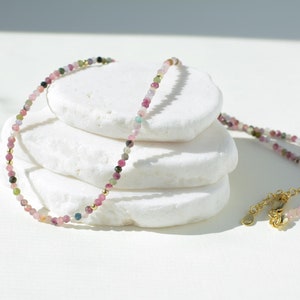 May include: Delicate gold necklace featuring multicolored tourmaline beads. The necklace is displayed on a stack of white stones, showcasing its dainty design and vibrant gemstone colors.  The stones appear to be pink, green, and dark brown. The necklace has a simple clasp closure.