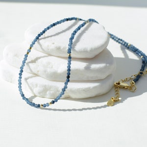 May include: A delicate blue gemstone necklace with gold accents. The necklace is made of small, round beads and has a simple, elegant design.