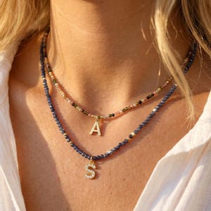 May include: A layered necklace set featuring two strands of small faceted beads. The shorter strand has multicolored beads with a gold letter A pendant. The longer strand has dark blue beads with a gold letter S pendant encrusted with clear stones.