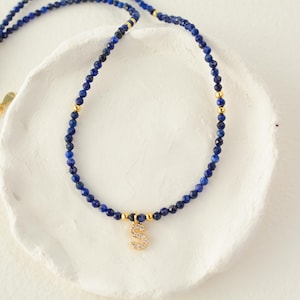 May include: A necklace featuring small, faceted deep blue lapis lazuli beads interspersed with tiny gold-toned metal spheres. A gold-toned letter S pendant encrusted with clear crystals hangs from the center of the beaded strand.
