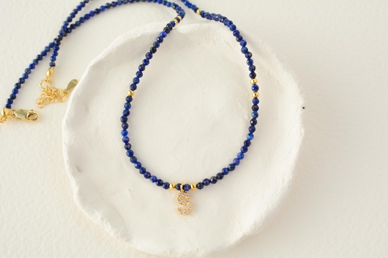 May include: A necklace featuring small, faceted deep blue lapis lazuli beads interspersed with tiny gold-toned metal spheres. A gold-toned letter S pendant encrusted with clear crystals hangs from the center of the beaded strand.
