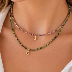 May include: Two layered necklaces featuring small, faceted, multi-colored gemstone beads. The shorter necklace has a gold-toned letter J pendant, while the longer necklace features a gold-toned letter G pendant, both with small embedded stones.