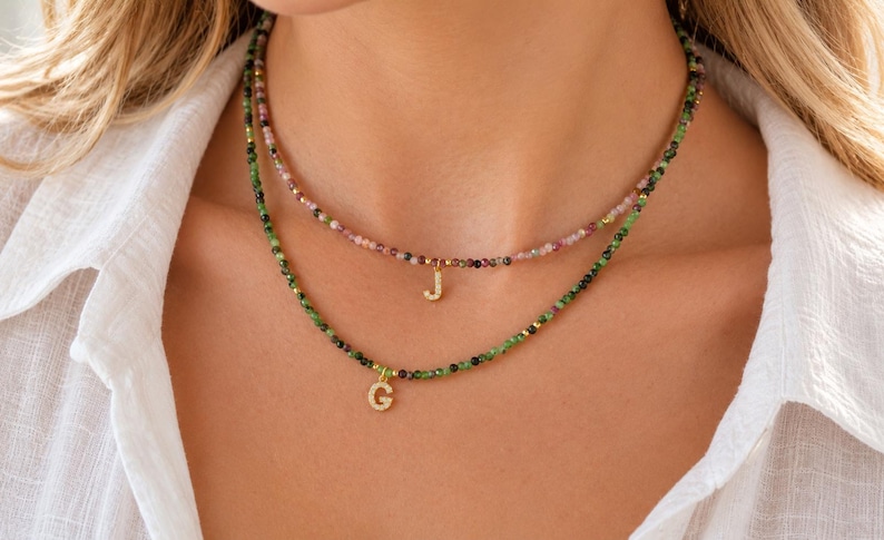 May include: Two layered necklaces featuring small, faceted, multi-colored gemstone beads. The shorter necklace has a gold-toned letter J pendant, while the longer necklace features a gold-toned letter G pendant, both with small embedded stones.