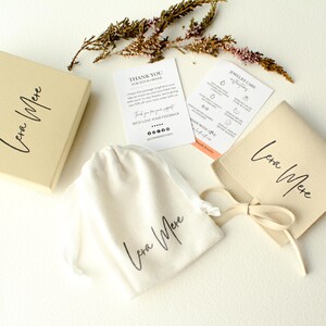 May include: A small, cream-colored pouch with a brown ribbon tied around it. The pouch has the words "Lena Mere" printed on it in black script. The pouch is sitting on a white surface next to a small card with the words "Jewelry Care Instructions" printed on it.