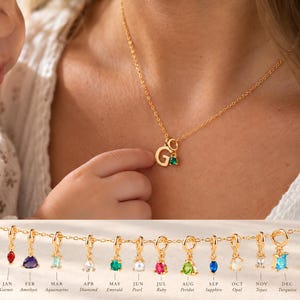 May include: A gold-tone chain necklace featuring a small letter G pendant and a tiny green gemstone charm. Below, a display shows twelve individual gold-tone charms, each with a different colored birthstone representing months January through December.