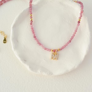May include: A beaded necklace featuring small pink gemstone beads, interspersed with tiny gold-toned metal spheres. The necklace includes a gold-toned letter M pendant encrusted with clear crystals and a gold-toned extender chain with an S925 tag.