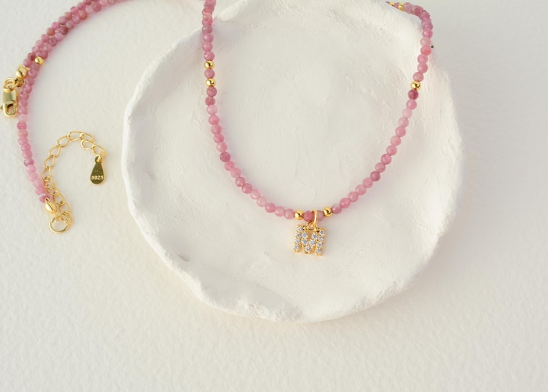 May include: A beaded necklace featuring small pink gemstone beads, interspersed with tiny gold-toned metal spheres. The necklace includes a gold-toned letter M pendant encrusted with clear crystals and a gold-toned extender chain with an S925 tag.