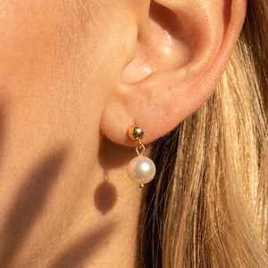 May include: A gold stud earring featuring a small polished gold sphere at the earlobe, connected to a dangling white spherical pearl. The earring is crafted with a simple metal link that allows the pearl to hang freely below the gold stud component.