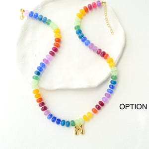 May include: A colorful beaded necklace with a rainbow gradient of beads. The necklace has a gold-colored clasp and a gold-colored initial pendant. The necklace is displayed on a white surface.