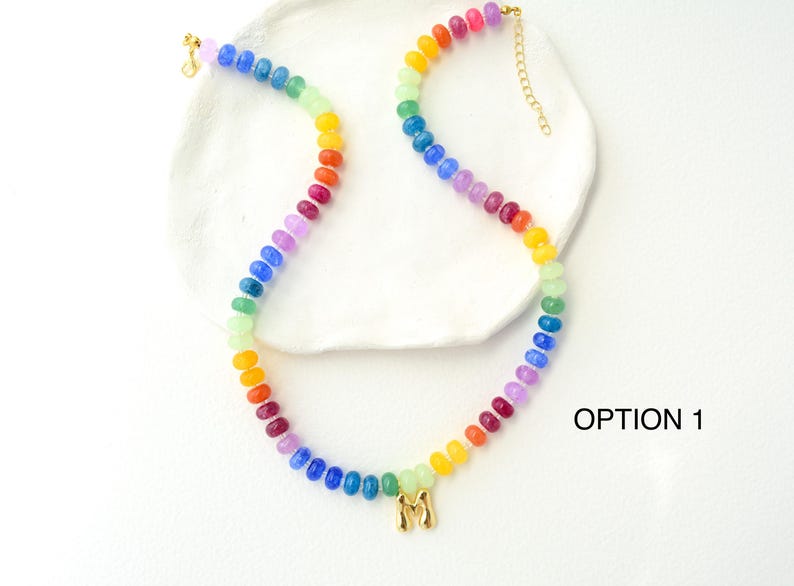 May include: A colorful beaded necklace with a rainbow gradient of beads. The necklace has a gold-colored clasp and a gold-colored initial pendant. The necklace is displayed on a white surface.