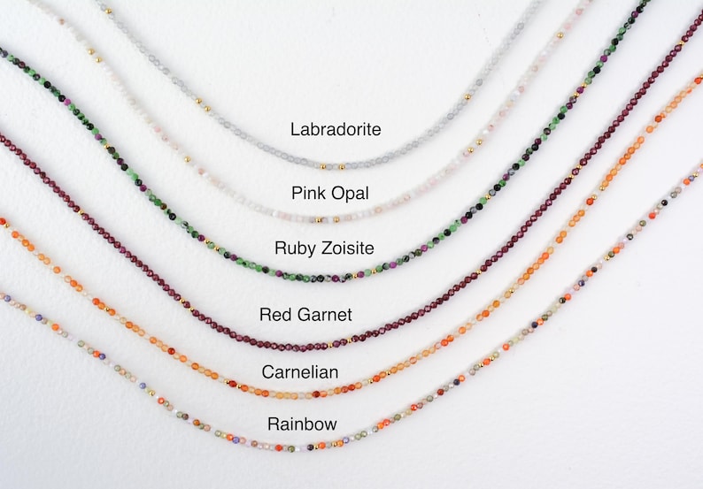 May include: Six delicate beaded necklaces arranged in parallel arcs, featuring Labradorite, Pink Opal, Ruby Zoisite, Red Garnet, Carnelian, and Rainbow gemstone beads, each accented with small gold-toned metal spacers throughout the strands.