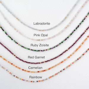 Puede incluir: Primer plano de siete collares con gemas de diferentes colores. Los collares est&aacute;n dispuestos en fila de arriba a abajo, siendo el collar superior el de color m&aacute;s claro y el inferior el de color m&aacute;s oscuro. Los collares est&aacute;n etiquetados con los nombres de las gemas: Labradorita, &Oacute;palo rosa, Zoisita rub&iacute;, Granate rojo, Cornalina y Arco&iacute;ris.