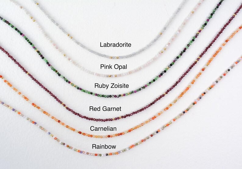 Puede incluir: Primer plano de siete collares con gemas de diferentes colores. Los collares est&aacute;n dispuestos en fila de arriba a abajo, siendo el collar superior el de color m&aacute;s claro y el inferior el de color m&aacute;s oscuro. Los collares est&aacute;n etiquetados con los nombres de las gemas: Labradorita, &Oacute;palo rosa, Zoisita rub&iacute;, Granate rojo, Cornalina y Arco&iacute;ris.