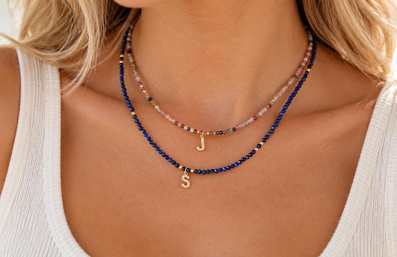 May include: Two layered beaded necklaces featuring small faceted stones. The top necklace has multicolored beads with a gold letter J pendant. The bottom necklace has deep blue beads with gold accents and a gold letter S pendant with small stones.