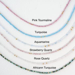 May include: A collection of delicate necklaces featuring various gemstones, including pink tourmaline, turquoise, aquamarine, strawberry quartz, rose quartz, and African turquoise. Each necklace is made with small beads and gold accents.