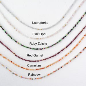 May include: A close-up of seven necklaces with different colored gemstones. The necklaces are arranged in a row from top to bottom, with the top necklace being the lightest color and the bottom necklace being the darkest color. The necklaces are labeled with the names of the gemstones: Labradorite, Pink Opal, Ruby Zoisite, Red Garnet, Carnelian, and Rainbow.