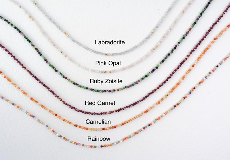 May include: A close-up of seven necklaces with different colored gemstones. The necklaces are arranged in a row from top to bottom, with the top necklace being the lightest color and the bottom necklace being the darkest color. The necklaces are labeled with the names of the gemstones: Labradorite, Pink Opal, Ruby Zoisite, Red Garnet, Carnelian, and Rainbow.