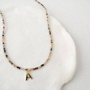 May include: A delicate necklace featuring small, multi-colored gemstone beads in shades of pink, green, and brown, accented with tiny gold-toned spacers and a single gold-toned letter A pendant hanging from the center of the strand.