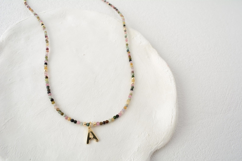 May include: A delicate necklace featuring small, multi-colored gemstone beads in shades of pink, green, and brown, accented with tiny gold-toned spacers and a single gold-toned letter A pendant hanging from the center of the strand.