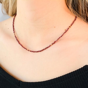 May include: A delicate necklace with small, dark red beads and gold accents. The necklace is worn by a person with a black shirt.