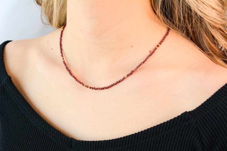 May include: A delicate necklace with small, dark red beads and gold accents. The necklace is worn by a person with a black shirt.