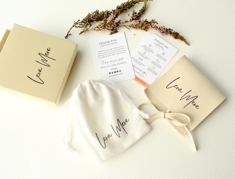 K&ouml;nnte beinhalten: Eine kleine, cremefarbene Tasche mit einem braunen Band, das um sie herum gebunden ist. Die Tasche tr&auml;gt die Aufschrift "Lena Mere" in schwarzer Schrift. Die Tasche liegt auf einer wei&szlig;en Oberfl&auml;che neben einer kleinen Karte mit der Aufschrift "Anleitung zur Schmuckpflege".