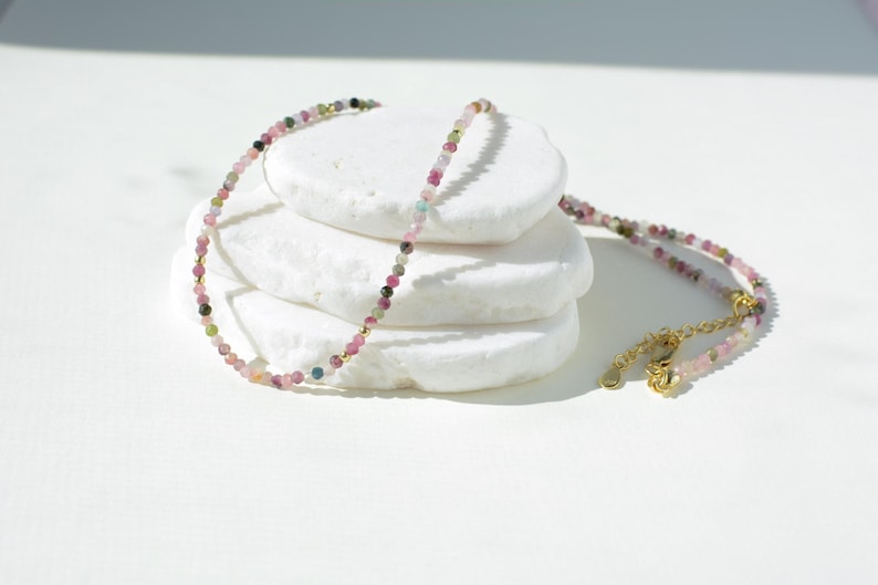 May include: Delicate gold necklace featuring multicolored tourmaline beads. The necklace is displayed on a stack of white stones, showcasing its dainty design and vibrant gemstone colors.  The stones appear to be pink, green, and dark brown. The necklace has a simple clasp closure.