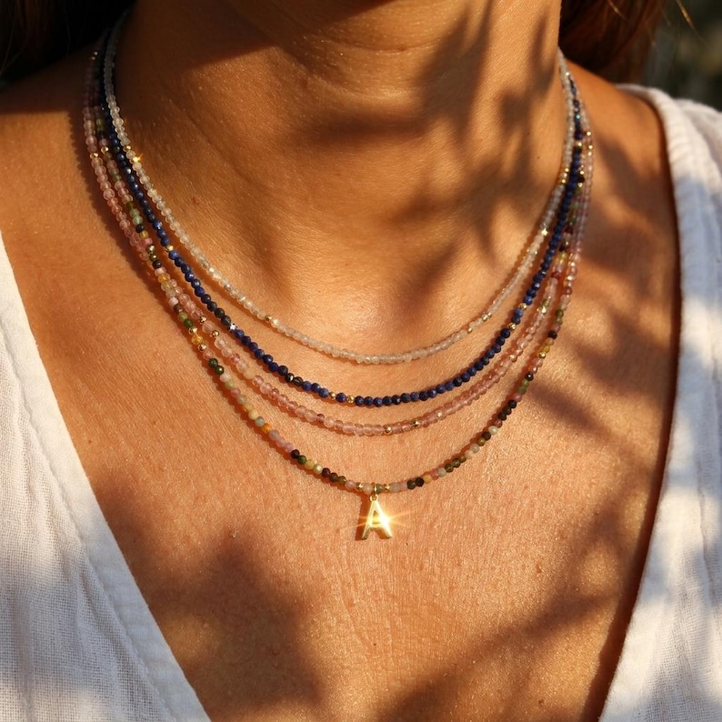 May include: A layered necklace set consisting of four strands of small, faceted, multi-colored gemstone beads. The strands feature shades of light pink, deep blue, and earth tones, accented with tiny gold-colored beads and a gold-colored letter A charm.