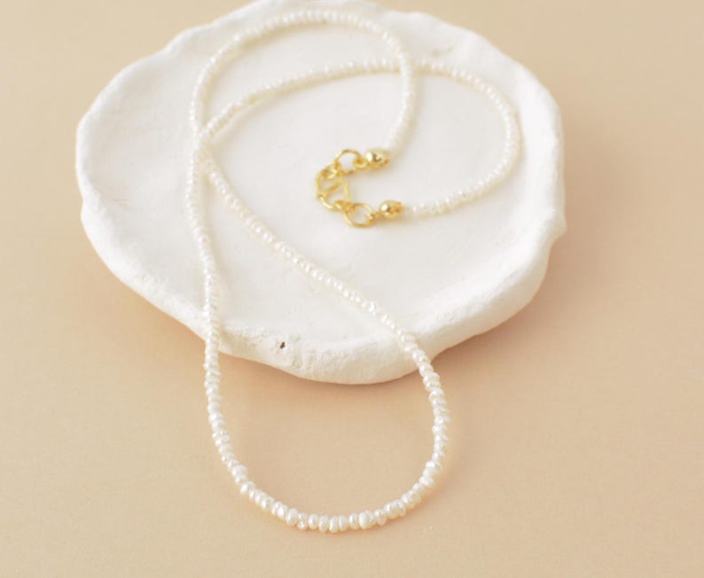 May include: A delicate white pearl necklace with a gold clasp. The necklace is displayed on a white ceramic dish.