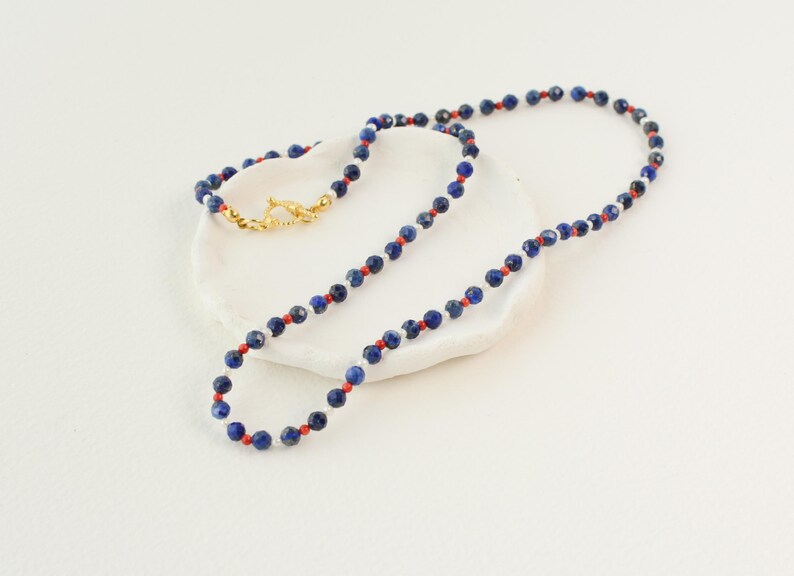 May include: A necklace made of blue, red, and white beads. The necklace is on a white surface.