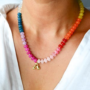 May include: A colorful beaded necklace with a gold pendant. The necklace features a gradient of colors, transitioning from blue to pink to orange and yellow. The gold pendant is a stylized letter "A".