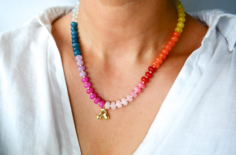 May include: A colorful beaded necklace with a gold pendant. The necklace features a gradient of colors, transitioning from blue to pink to orange and yellow. The gold pendant is a stylized letter "A".