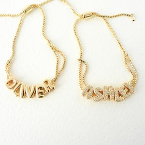 May include: Two gold-colored bracelets with adjustable chains. One bracelet spells "OLIVER" in gold block letters. The other bracelet spells "ASHLEY" in gold letters encrusted with small, clear crystals. Both bracelets are on a white background.