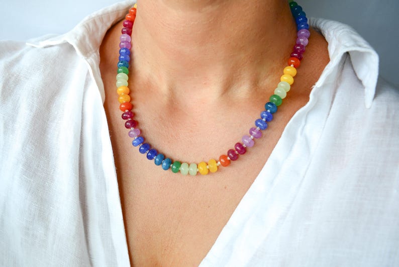 May include: A colorful beaded necklace with a rainbow of colors, including red, orange, yellow, green, blue, and purple. The necklace is worn against a white shirt.
