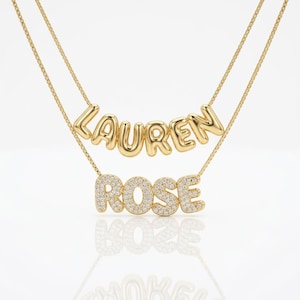 May include: A layered gold necklace featuring two chains. The top chain displays the name LAUREN in smooth, polished gold balloon-style letters. The bottom chain features the name ROSE in gold letters encrusted with small clear stones.
