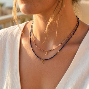 May include: A layered necklace set featuring two strands of small, faceted beads. The shorter strand has multicolored beads with a gold-toned letter J charm, while the longer strand has deep blue beads with a gold-toned letter S charm.