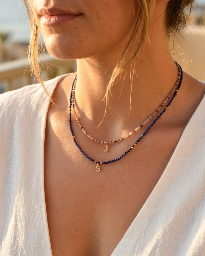 May include: A layered necklace set featuring two strands of small, faceted beads. The shorter strand has multicolored beads with a gold-toned letter J charm, while the longer strand has deep blue beads with a gold-toned letter S charm.