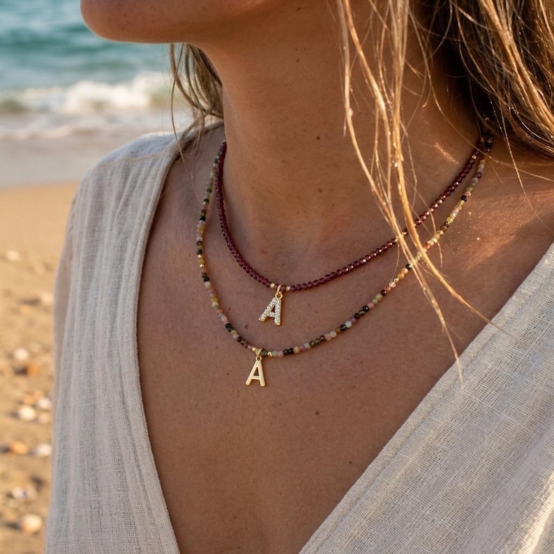 May include: A layered necklace set featuring two strands of small beads. The top strand consists of dark red beads with a gold-toned letter A pendant. The bottom strand features multi-colored beads with a gold-toned letter A pendant.