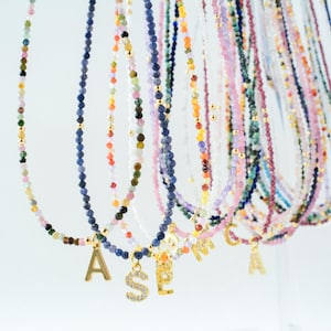 May include: A collection of delicate beaded necklaces featuring small, multi-colored gemstone beads and gold-toned spacers. Each necklace is accented with a single gold-toned letter charm, some of which are embellished with small clear stones.