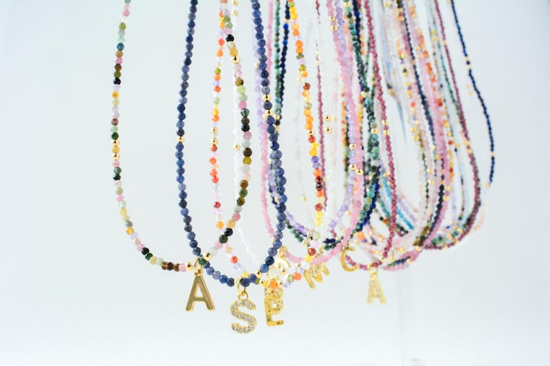 May include: A collection of delicate beaded necklaces featuring small, multi-colored gemstone beads and gold-toned spacers. Each necklace is accented with a single gold-toned letter charm, some of which are embellished with small clear stones.