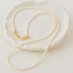 May include: A delicate white pearl necklace with a gold clasp. The necklace is displayed on a white surface.
