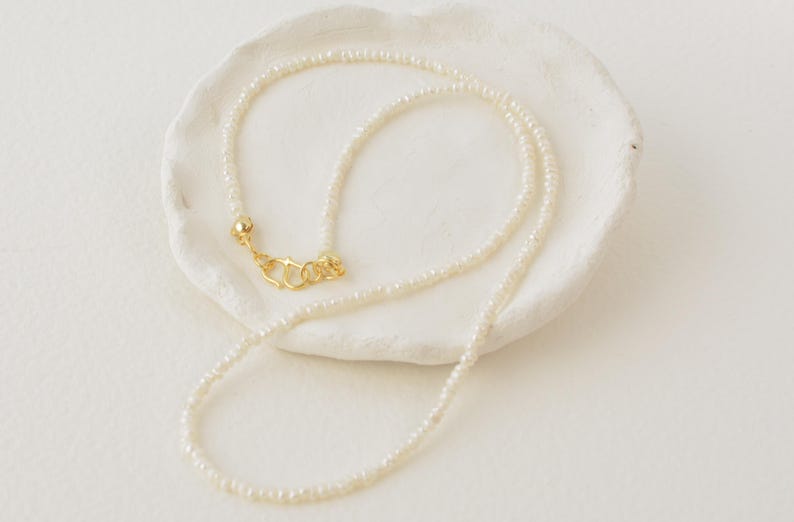 May include: A delicate white pearl necklace with a gold clasp. The necklace is displayed on a white surface.