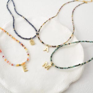 May include: Four beaded necklaces featuring small, multi-colored faceted stones and gold-toned metal accents. Each necklace includes a single gold-toned letter charm, specifically the letters E, S, A, and M, hanging from the center.