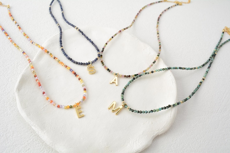May include: Four beaded necklaces featuring small, multi-colored faceted stones and gold-toned metal accents. Each necklace includes a single gold-toned letter charm, specifically the letters E, S, A, and M, hanging from the center.