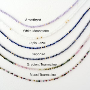 May include: A close-up of six different necklaces made with gemstone beads. The necklaces are arranged in a row from top to bottom, with the top necklace being amethyst, followed by white moonstone, lapis lazuli, sapphire, gradient tourmaline, and mixed tourmaline.