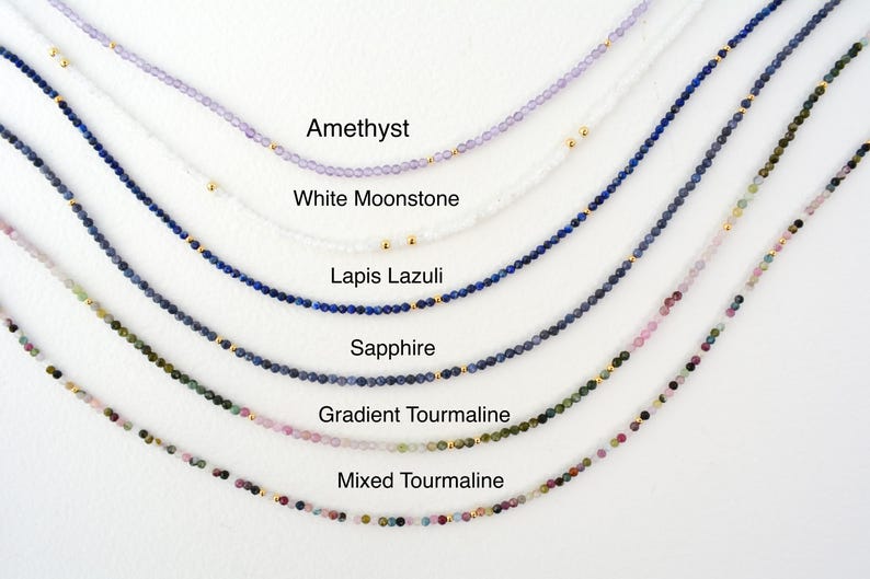 May include: A close-up of six different necklaces made with gemstone beads. The necklaces are arranged in a row from top to bottom, with the top necklace being amethyst, followed by white moonstone, lapis lazuli, sapphire, gradient tourmaline, and mixed tourmaline.