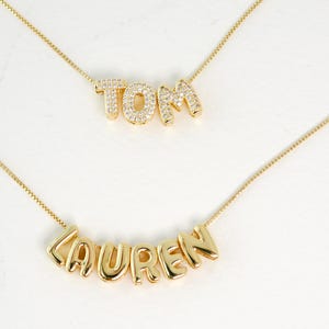 May include: Two gold necklaces with the names "TOM" and "LAUREN" spelled out in gold letters with small clear stones. The necklaces are on a white background.
