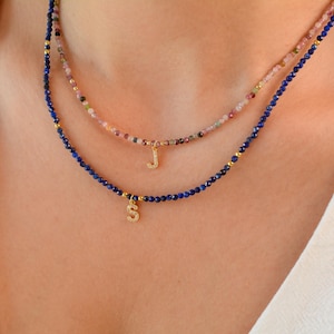 May include: Two layered necklaces with gold initial charms. The top necklace is made of small pink and white beads. The bottom necklace is made of dark blue beads.